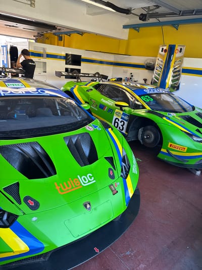 This image captures two green and yellow racing cars, presumably Lamborghini Huracáns, inside a garage or pit area. The car in the foreground is partially visible, showcasing its aerodynamic design with a prominent front splitter. Its livery includes a red "Loc" logo, the Lamborghini emblem, and a "BURGERS" sponsor decal. The other car, numbered 63, is positioned behind it. This car bears the "IRELLI" tire logo. Mechanics are present, one of whom is kneeling and working on equipment in the background near a racing team support vehicle. The environment appears to be a pit area, with the colors yellow, white, and blue. The image implies the location could be the Autodromo Vallelunga Piero Taruffi circuit near Campagnano di Roma, Italy, considering the presence of racing cars and the professional racing atmosphere.