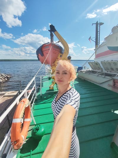 In a selfie taken on the deck of a vessel, a woman with blonde hair and red lipstick poses in front of a life raft and the open sea under a bright blue sky with scattered clouds. She wears a black and white striped dress. The vessel's deck is painted green, and to her left, a life preserver is mounted on the railing. In the background, the water stretches to a tree-lined shore. The image appears to be taken in Kvartsitny, Russia.