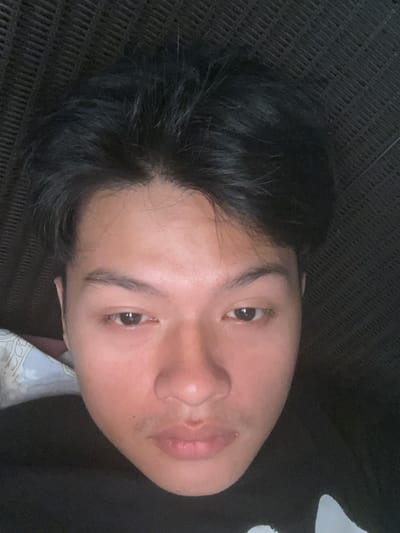 This is a close-up, overhead, selfie-style photograph of a young East Asian male lying down. He has short, dark, messy hair and is wearing a black t-shirt. The background consists of a dark, woven textured surface, possibly a piece of furniture. The lighting is dim, creating a somewhat shadowy effect. The image is taken from a low angle, focusing on the man's face, with his head tilted slightly. The man's eyes are open and looking slightly off-camera. His skin tone is fair. There is a hint of a patterned fabric on the left side of the image, possibly a pillow or blanket. The scene appears to be indoors in a residential setting within the city of Serdang Kulon, Indonesia.