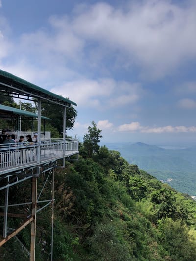 A metal walkway with a green roof extends from the left side of the frame towards the right, elevated above a steep, densely vegetated mountainside. Several people are standing and sitting on the walkway, which is enclosed by white metal railings. The walkway appears to be part of a tourist attraction or observation deck. The background is a panoramic view of lush green mountains and valleys under a partly cloudy blue sky. A body of water can be seen in the distance, nestled among the rolling hills. The scene suggests a scenic overlook in a mountainous region, likely a popular spot for visitors to enjoy the natural landscape. The location is identified as being in Nam Hải, Vietnam.