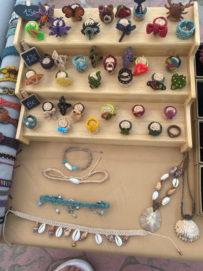 The image shows a close-up of a market stall displaying handmade macrame jewelry and decorative items. The stall features a multi-tiered wooden display filled with various small, colorful macrame creations, many of which are mounted on cork stoppers. These items include butterflies, dragonflies, spiders, hearts, fruits, and abstract shapes. Some items are priced with small chalkboard tags. In front of the display, several bracelets and necklaces are laid out on a tan fabric surface. These include macrame bracelets with beads and shells, a turquoise macrame bracelet with a starfish charm, and necklaces adorned with seashells. The background is slightly blurred, showing a glimpse of other stalls or merchandise, suggesting a bustling outdoor market environment. The scene is consistent with a vendor setting up or displaying goods at a beachside market in Playa Del Sol-Villacana, Spain, given the style of the jewelry and the potential for a coastal location. There are no visible people in the frame.