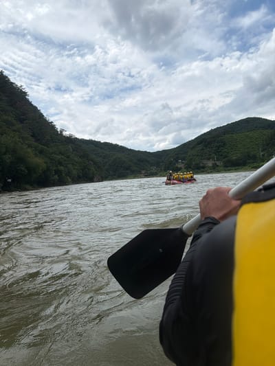 In this perspective shot, a person is holding a paddle, their arm and part of their yellow life vest visible in the foreground. The paddle is poised over the rippling surface of a wide river. In the middle ground, a red raft carrying several individuals wearing yellow life vests is moving down the river. The riverbanks are lined with lush green, tree-covered hills, suggesting a rural or natural setting. The sky above is filled with a mix of dramatic, grey clouds and patches of blue, indicating an overcast or changing weather condition. The scene depicts a rafting activity, likely taking place on the river that flows through Seo-myeon, South Korea, known for its scenic natural landscapes and outdoor recreational opportunities.