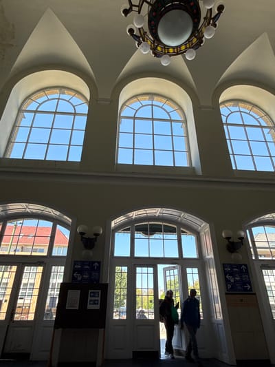 This media file captures the interior of a grand, high-ceilinged building, identified as a transport hub, likely a train or bus station in Szeged, Hungary. The image is oriented vertically, showing the ceiling and upper walls on the left, and a wall with windows and a doorway on the right.
The architecture is notable for its arched and curved ceilings and numerous large windows. On the left side of the image (which would be the ceiling if correctly rotated), three large, semi-circular arched windows illuminate the space with natural light, showcasing a clear blue sky. A large, ornate chandelier with multiple white globe lights hangs prominently from the ceiling.
On the right side of the image (a wall), a series of rectangular and arched windows, along with glass doors, provides further illumination and views of the exterior. Through these windows, a bright, clear blue sky, parts of other buildings (including red brick and yellow/beige structures), and green trees are visible, indicating a sunny daytime setting.
Two individuals are silhouetted against the bright light from the doorway on the right, appearing to walk or stand near an entrance/exit. One person wears a blue jacket and dark pants, while the other wears a darker top, lighter pants, and appears to carry a green bag.
Mounted on the wall on the right is a blue informational sign with white pictograms and Hungarian text. Visible text includes "Post", "Taxi", "Poggyász" (luggage, likely indicating a train station), "Autóbusz" (bus), "Kerékpár" (bicycle), and "Parkolás" (parking), accompanied by directional arrows. Below this, a dark board contains additional, partially obscured information. Two smaller, wall-mounted light fixtures with two globe lights each are also visible on this wall.