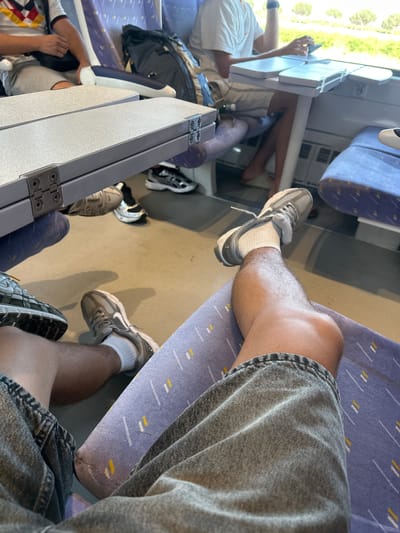 The image shows the interior of a train car. In the foreground, a person's legs are visible, clad in denim shorts and grey sneakers with white socks. The person's right leg is bent at the knee, with the foot resting on the seat. The train car features blue seats with a patterned design. Several other people are visible in the background, also seated, with some luggage present. Fold-down tables are attached to the backs of the seats. The scene appears to be set during daytime, with natural light coming through the windows, suggesting the train is traveling through a landscape with some trees visible outside. The image does not provide any specific visual cues to pinpoint the location within Salses-le-Château, France.