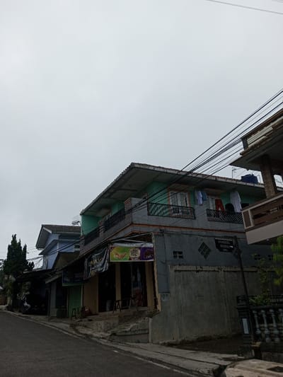 This scene is located in the city of Ambakirano, Madagascar. The image captures an urban street under an overcast, light grey sky. An asphalt road occupies the lower-left foreground, gently inclining upwards towards the left, bordered by a concrete sidewalk on the right.
A two-story building dominates the right side of the frame. Its upper floor features a light teal facade with a black metal balcony railing. Several items of clothing, including light-colored shirts, a pair of red pants, and dark shirts, are hung on lines over the balcony for drying. Two sets of double doors or large windows open onto this balcony. The ground floor of this building is constructed of grey concrete. Two commercial banners are attached to the ground floor; one is a dark, partially obscured banner, and the other is a lighter banner depicting food (possibly a pastry) with text that includes "Jualan." Below these banners, an entrance to a shop is visible, with two light-colored stools placed on the steps. To the right of the shop entrance, a large, vertical, lighter grey panel, possibly a garage door, is present. A signpost next to this panel displays the text "BENGKEL MOBIL" and "OBL."
Numerous dark utility cables traverse the upper middle section of the image, running horizontally in front of and above the central building. A blue plastic water tank is visible on the roof of an adjacent building to the far right.
To the left, another multi-story structure is partially visible behind the central building and a utility pole. This building has a light blue upper facade and a dark-tiled roof. Further down the street on the left, additional structures are interspersed with dense green foliage, including a tall, dark green coniferous tree. No people, vehicles, or animals are visible within the frame.