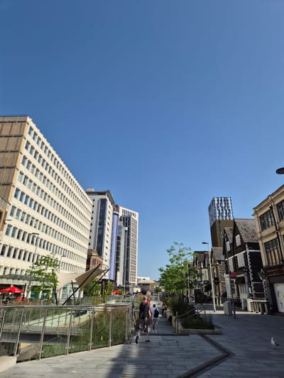 The image depicts a street scene in Cardiff, United Kingdom, characterized by a mix of modern and traditional architecture. On the right side of the image, pedestrians walk along a paved sidewalk, adjacent to a glass-encased garden or planting area. On the left side, tall modern buildings with repetitive window patterns rise high, contrasted by the older, more ornate buildings on the opposite side. The sky above is clear and blue, suggesting a sunny day. People are seen walking, suggesting a lively urban environment.