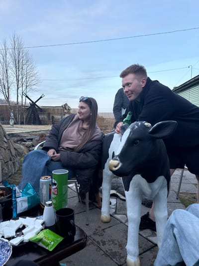 The image shows two individuals outside with a black and white cow statue. One individual is standing, leaning towards the cow statue, holding a green glass bottle. The other person is seated, looking down. There are Pringles, a drink can, food packets, and white foam pieces on a table or surface. The environment includes bare trees, a fence, and a building. There is no definitive evidence to confirm the location as Sukhobuzimskoye, Russia.
