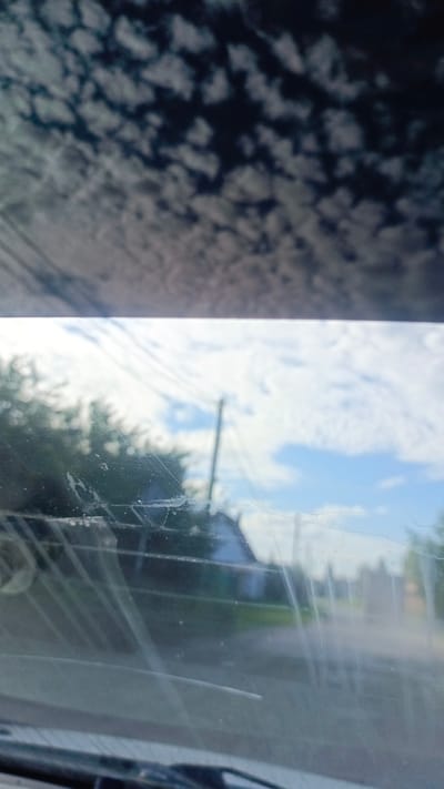 The image shows a view through a slightly dirty windshield. The upper portion of the image, reflecting the sky, displays scattered clouds against a dark sky. The lower portion of the image, showing the outside scene, is out of focus and features a bright sky with scattered clouds, trees on the left, a utility pole with wires, and a building in the distance. The windshield itself shows streaks and smudges. The scene appears to be a rural or suburban area, possibly on the outskirts of a city. Given the location, the image was likely taken in the city of Zuy, Russia.