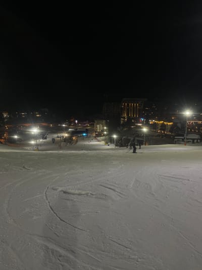 The image displays a night scene at a ski resort in Jahorina, Bosnia and Herzegovina. The foreground and midground feature a snow-covered ski slope, marked extensively with ski tracks. In the midground-right, a lone individual on skis is discernible, oriented downhill. Further down the slope, numerous smaller figures are visible, engaged in skiing or snowboarding activities, concentrated towards the base of the visible incline. Artificial illumination from streetlights and floodlights casts light across the slope and the distant resort village. The background reveals a cluster of illuminated buildings, including a prominent multi-story structure on the right exhibiting vertical architectural lighting. Elements of ski lift infrastructure are present in the midground-left. Construction cranes are visible among the background buildings, indicating ongoing development. The upper section of the image consists of a dark night sky.