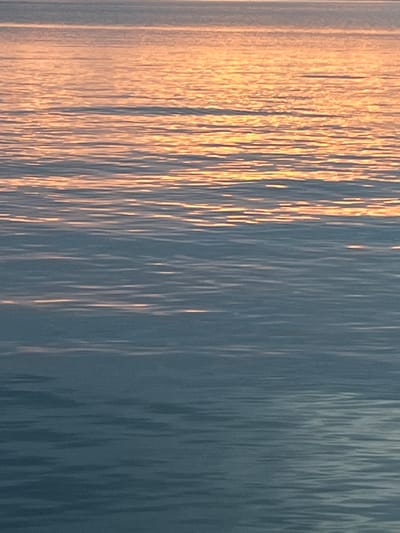 The image displays a close-up, horizontal view of a body of water, identified as Lake Geneva in Montreux, Switzerland. The upper two-thirds of the frame are dominated by warm, golden-orange light reflections stretched horizontally across the water's surface, indicative of a low sun angle (either sunrise or sunset). The lower third transitions into cooler, muted blue-grey tones, also exhibiting horizontal streaks of reflected light. The water surface shows consistent, subtle ripples and elongated patterns formed by the interaction of light and waves. No people, vessels, or other identifiable objects are present within the frame. The scene is an abstract representation of water and ambient light conditions.