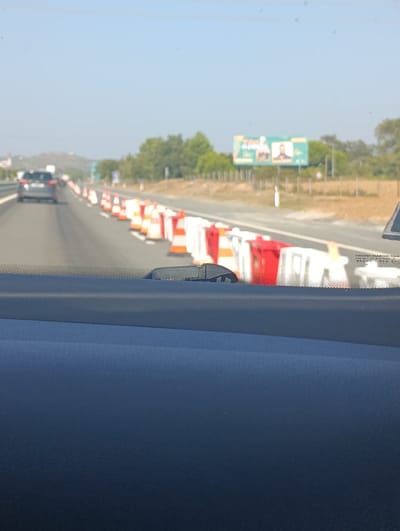 The image shows a view from inside a vehicle, looking forward onto a highway in Santiago do Cacém, Portugal. A line of orange and white traffic cones and red and white barriers are visible on the right side of the road, indicating road construction or lane closure. A dark-colored car is in the left lane ahead of the viewpoint. In the background, there are trees, a billboard, and a hillside. The sky is clear and blue.