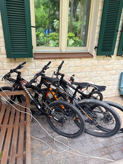 Four bicycles are parked in front of a brick building with green shutters. The bicycles are lined up next to each other, with the front wheel of the first bicycle resting on a wooden bench. The bicycles are black and orange and appear to be mountain bikes. The building has a large window that reflects the surrounding greenery. The scene appears to be outdoors, possibly in a residential area. The city of Sosny, Russia is known for its natural beauty and is often visited by tourists.