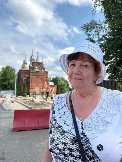 In Chudovo, Russia, an elderly woman with auburn hair, wearing a white sun hat and a white blouse with a black and white patterned V-neck, stands in the foreground, smiling. She is adorned with a gold chain necklace featuring a cross pendant. Behind her, a red brick church with silver domes and crosses is visible, appearing to be undergoing construction or renovation, with barriers and scaffolding in the surrounding area. Trees and a cloudy sky form the backdrop.