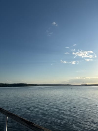 The image depicts a serene body of water, likely a lake or bay, with a wooden railing in the foreground. In the distance, a tree-covered shoreline is visible, followed by a hazy horizon. On the right side of the horizon, industrial structures, including tall chimneys, are present, suggesting a nearby industrial area. The sky is a clear blue with scattered white clouds, and the lighting indicates it is either early morning or late afternoon. The water is calm, with gentle ripples reflecting the sky. No people or animals are visible in the image. The location is identified as Kazashko, Bulgaria.