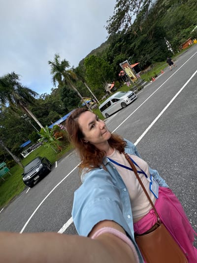 A woman with shoulder-length auburn hair and a light-colored tank top, blue button-down shirt, and bright pink skirt is taking a selfie on a paved road in Tambon Wang Prachan, Thailand. She is wearing a brown crossbody bag and a blue lanyard with an identification card. The road has white lane markings and is bordered by a grassy shoulder. In the background, there are palm trees, lush green foliage, a dark SUV parked on the side of the road, and a white van further down the road. The sky is overcast. A tall banner with religious imagery stands near the road, and there are various small buildings and signs in the distance. The overall impression is of a tropical roadside environment.