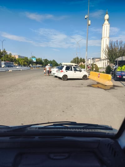 The image depicts an outdoor scene in Buz, Uzbekistan, on a clear day with a blue sky. In the foreground, the interior of a vehicle is visible, including the dashboard and windshield wipers. The road in front of the vehicle is paved with asphalt and shows some wear and tear, with a puddle of water and some debris present.
Several vehicles are parked or driving on the road. Two white cars are prominent, one of which appears to be parked somewhat haphazardly near a large, yellow concrete barrier. A blue truck with a canopy, loaded with watermelons, is parked on the right side of the road. A man and a child are walking away from the camera on the left side of the road, heading towards the white cars.
In the background, a tall, ornate minaret of a mosque stands against the sky, indicating a religious or cultural landmark. Trees line the sides of the road, and other buildings and vehicles are visible in the distance. A security camera is mounted on a pole near the minaret. The overall impression is a street scene in a city, with everyday activities taking place.