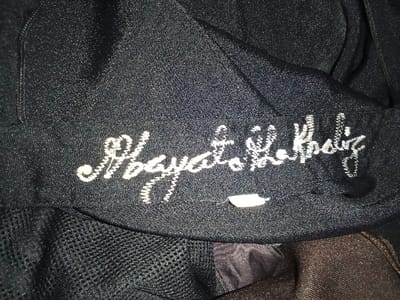 The image displays a close-up, top-down view of a dark, textured textile item, likely synthetic fabric of a garment or accessory. White, stylized cursive embroidery reading "Kgaya La Kposiz" is centrally featured. A small, partially obscured white fabric tag is visible directly beneath the embroidered text. To the lower left, a section of black mesh fabric is present. To the lower right, a crumpled area of dark brown fabric is visible. The lighting is dim and uneven, accentuating the fabric's texture. No individuals, actions, or broader environmental context are discernible. The specific location within Umsobomvu Local Municipality, South Africa, cannot be determined from the visual information provided in the image.