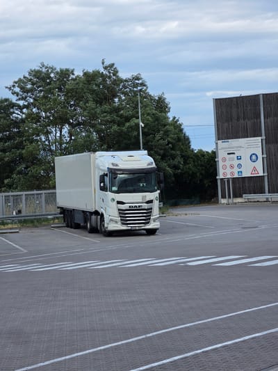 A white DAF truck is parked in a paved lot with white lane markings. The truck has a refrigerated trailer. To the left of the truck, there are green trees and a blue sky with white clouds. Behind the truck and to the right, there is a tall, dark-colored fence with a sign containing multiple pictograms and text. The text on the sign includes "ACHTUNG FUSSGÄNGER! LOOK OUT FOR PEDESTRIANS!" and "NAGEL-GROUP". Other visible text includes the truck's license plate "MOB 592" and the brand name "DAF" on the front of the truck. The scene appears to be outdoors during the daytime with fair weather. The parking lot suggests a commercial or industrial area, possibly a logistics hub or a truck stop. There are no people visible in the image.