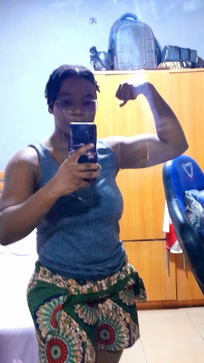 A person stands in front of a mirror, holding a mobile phone to take a picture. The person is wearing a grey tank top and a patterned skirt. They have their arm raised and flexed. Behind the person, there is a wooden cabinet with items on top, including a blue cushion and a bag. The location is indicated as Umuigbekwo, Nigeria, although this is not visually verifiable.