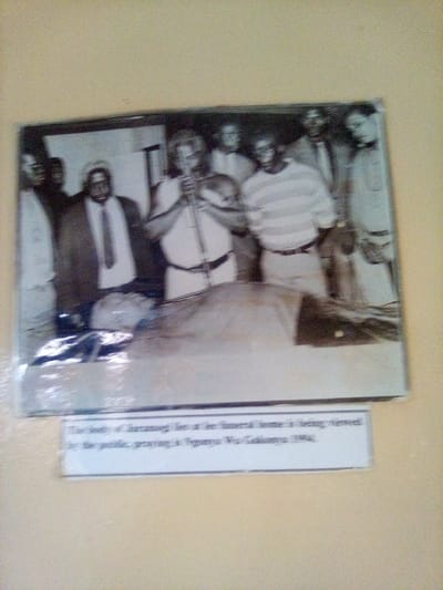 A black and white photograph, exhibiting some grain and low contrast, is adhered to a light-colored, possibly pale yellow, wall. The photograph depicts an indoor scene with eight dark-skinned individuals. In the foreground, a deceased individual lies supine, partially covered by a light-colored sheet, with their head positioned towards the left of the frame. Seven adult males stand around the deceased, observing the body. One man, centrally located behind the deceased, wears a light-colored top and a dark belt, holding a long, thin, light-colored staff or pipe vertically with both hands. To his left, another man wears a dark suit jacket, white shirt, and dark tie. To the right, a man wears a horizontally striped long-sleeved shirt and light-colored trousers. The remaining individuals are less distinct but appear to be wearing shirts and trousers. A white rectangular label with black text is affixed to the wall directly below the photograph, stating: "The body of Jaramogi lies at a funeral home is being viewed by the public, praying is Yganya Wu Gokonya 1984." This entire display is located in South Sakwa ward, Kenya.