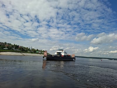 This is a wide shot of the Volga River in Verkhniy Uslon, Russia, on a cloudy day. In the foreground, the dark, choppy water of the river dominates the lower half of the frame. In the midground, a dark ferry boat with a white superstructure is moving from left to right across the river. The ferry has a license plate that reads "CH-18". On the left bank of the river, a village with houses and green trees sits on a hillside. The village is protected by a concrete embankment. In the distance, the river extends to the horizon, flanked by a dense line of trees. The sky is filled with a mix of white and gray clouds against a bright blue sky. Two red buoys are visible in the water in the distance.
