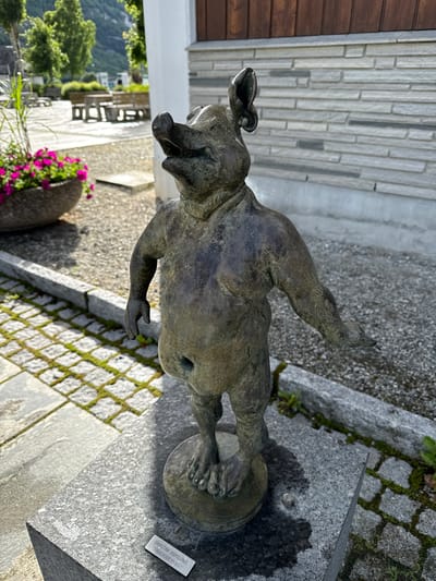 A bronze statue of a pig-like creature with human-like legs stands on a granite pedestal in what appears to be a public outdoor space in Eidfjord, Norway. The statue depicts a figure with a snout, pig ears, and a distended belly, with its arms outstretched and its mouth open as if speaking or singing. The pedestal is labeled with a small metal plaque that reads "CHRISTINE ASPELUND 'GRISEHODETROLL'".
The environment suggests a town square or park. Behind the statue, there is a building with a textured grey stone facade and dark wooden accents. To the left of the statue, a large stone planter holds vibrant pink flowers, and beyond that, trees and distant mountains are visible. The ground is paved with cobblestones and gravel. The lighting indicates it is daytime, with sunlight casting shadows.