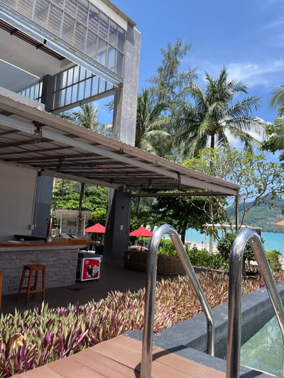 This image captures a resort pool area in Ban Bang Thao, Thailand, on a sunny day. The foreground features a swimming pool with metallic handrails, leading into the frame. Adjacent to the pool is a wooden deck, bordered by a dense planting of purple and green foliage. Beyond the plants, a tiled bar area is visible, complete with a wooden stool and an ice cream freezer. In the mid-ground, red umbrellas dot the landscape, hinting at lounging areas. Palm trees and lush vegetation frame the scene, with a tranquil turquoise ocean and a distant hillside visible in the background. The sky is a clear blue with scattered white clouds.
