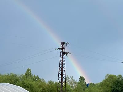 The image captures a scenic view in Balgarchevo, Bulgaria, featuring a large electricity pylon against a blue-grey sky with a rainbow. A tall, dark brown metal lattice pylon stands prominently near the center, supporting multiple power lines with white insulators that extend horizontally across the frame.

A faint but distinct rainbow arches diagonally across the sky, passing directly behind the pylon. Its colors are visible, stretching from the upper left, behind the pylon, and descending towards the lower right corner of the image. The rainbow appears to originate or terminate near the ground level amidst a line of lush green trees that form the horizon.

Below the power lines and pylon, a thick band of deciduous trees with vibrant green foliage fills the lower portion of the frame. In the bottom right corner, a small, distant figure in a blue shirt can be seen partially obscured by the trees, standing near where the rainbow meets the ground. On the far left, a portion of a white, curved, possibly corrugated structure (like a roof or large tent) is visible in the foreground. Small, white objects, potentially insulators or birds, are seen hanging from the power lines to the left of the pylon.

The sky is a soft, uniform blue-grey, suggesting either an overcast day or a recently cleared sky after rain, which would facilitate the formation of the rainbow. The time of day appears to be daylight, though specific indicators like strong shadows are absent. No discernible text is visible within the image. The scene offers a blend of natural wonder and industrial presence.