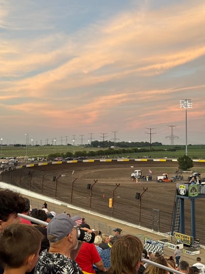 This is a medium-angle shot of the Dirt Oval at Route 66 Raceway in Joliet, Illinois, during a sunset or dusk event. The sky displays soft, peach and blue hues from the setting sun, with wispy clouds. In the foreground, a crowd of spectators are seated in bleachers, some holding drinks and wearing baseball caps. A man in a black and white patterned shirt and a gray cap with an American flag on it is visible in the mid-ground, holding a black can.
The race track, a dirt oval, occupies the central and lower portions of the image. Several vehicles, including trucks and what appears to be a tow truck, are positioned on the track and in the pit area. A tall light tower with bright stadium lights is illuminated on the right side of the track. In the background, a line of power transmission towers stretches across a flat, green landscape. The Victory Lane structure, adorned with trophies and signage for "Dirt Oval Route 66" and "Monster Truck," is prominent on the right side of the image, near the finish line. The overall scene depicts a racing event in progress or concluding, with a lively atmosphere among the spectators.