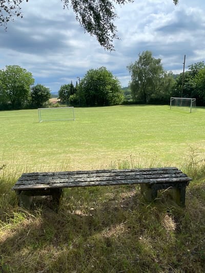 The image captures an outdoor scene featuring a weathered wooden bench in the foreground, positioned amidst overgrown grass. Behind the bench, a well-maintained green field, presumably a sports field, stretches into the background, marked by two soccer goals. Lush trees and vegetation frame the field, with a hilly landscape visible in the distance. The sky is overcast with dramatic clouds. The location is identified as a sports field in the city of Olberode, Germany.