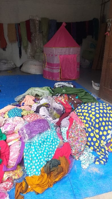 The image depicts an indoor scene in Cot Madi, Indonesia, with the floor largely covered by a blue tarp. A substantial, multi-colored pile of textile items, including various fabrics and what appear to be bundled clothing, occupies the foreground and midground. This pile exhibits a range of patterns such as polka dots, floral prints, and stripes, alongside solid colors, with some items contained within clear plastic packaging. In the background, a pink children's play tent, featuring a pointed roof, gold trim, and a crown motif, stands upright. Behind the tent, along a light-colored wall, various rolls of fabric or textiles are visible, some wrapped in clear plastic. Several large, off-white sacks are situated on the floor near the wall. To the right, a wooden door and doorframe are partially visible. A small, light-colored bird is present on the floor near the doorframe. No individuals are depicted, and no direct actions or interactions are occurring.