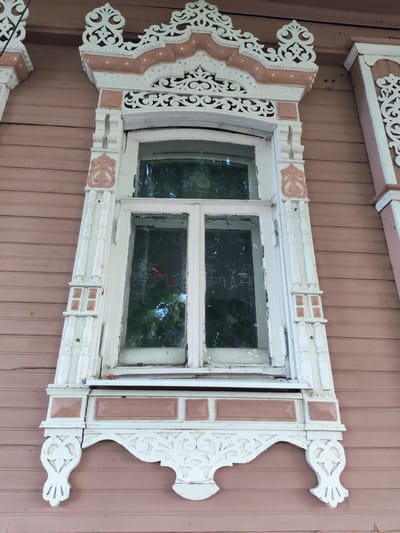 The image shows a close-up view of a decorative window frame on a wooden house. The house siding is painted a muted pinkish-brown. The window frame itself is white with carved details, featuring ornate scrollwork and geometric patterns. There are two panes of glass in the window, through which foliage and possibly flowers can be seen. The window is set within a larger decorative architectural element that extends above and to the sides of the window. This element is also predominantly white with pinkish-brown accents. The overall impression is one of traditional, possibly folk, architecture. The location provided is Chelganovo, Russia.