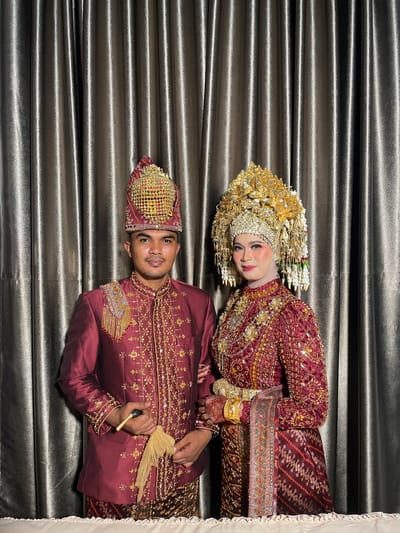 A couple poses for a portrait in traditional wedding attire against a backdrop of dark, pleated curtains. The man, on the left, wears a maroon jacket adorned with gold embroidery and a tall, ornate headdress. He holds a small fan-like object in his right hand. The woman, on the right, wears a similarly ornate maroon gown and a large, intricate gold headdress with hanging elements. She has noticeable makeup and jewelry, including gold bracelets. The background is a dark, metallic-looking curtain that is softly lit, creating vertical lines and highlights. The setting appears to be an indoor event, possibly a wedding reception hall, in the city of Tuha Peudaya, Indonesia.