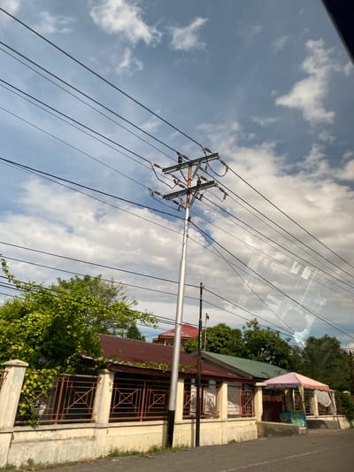 The image shows a daytime scene in Lampeudeu Baroh, Indonesia, featuring a utility pole with multiple power lines extending across a cloudy sky. The pole is located near a building with a brown roof and off-white walls; windows are covered by metal grills. Green foliage is visible around the base of the building and adjacent to the pole. The edge of a paved road is seen to the right of the building.