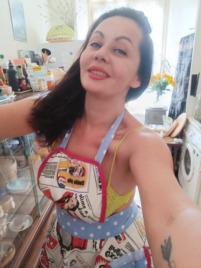 A medium shot captures a woman with dark hair and red lipstick taking a selfie in a kitchen. She is wearing a yellow bikini top and a blue polka-dot apron with a retro pattern. The apron features images of women and text, some of which is reversed. The background shows kitchen cabinets, shelves stocked with various items including bottles and containers, a washing machine, and a window with a curtain. The woman is smiling and looking at the camera, with her right arm extended to take the photo. The lighting appears to be natural, coming from the window.