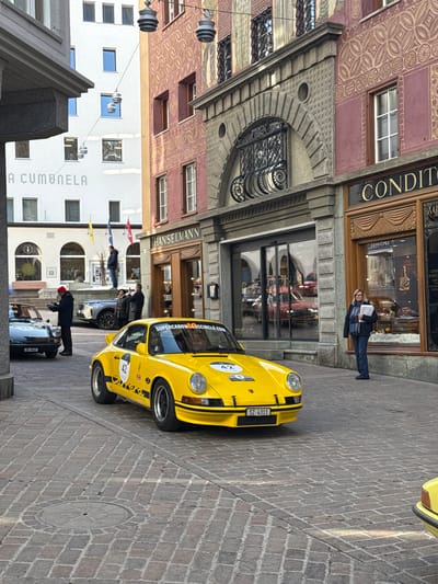 The scene depicts a street in Sankt Moritz, Switzerland, dominated by a bright yellow vintage Porsche 911, car number 42, centrally positioned on a cobblestone surface. This vehicle displays black number "42" decals on its hood and doors, "SUPERCARSOCIETY.COM" branding, and a Swiss license plate "SZ-4311."

To the left of the yellow Porsche, a dark grey vintage Porsche 911, car number 43, with license plate "ZG-5047," is parked. A man in a red beanie and dark coat stands beside it, holding and examining paperwork. Further back, a silver Cadillac XT6 SUV is visible, with two individuals partially obscured behind it. A portion of another yellow vehicle is present in the lower right foreground.

On the right side of the street, a woman wearing a dark coat and blue scarf stands on the pavement, holding a stack of white papers, facing towards the yellow Porsche. She is positioned in front of the Hanselmann building, which features a distinct maroon and gold patterned facade, ornate window grilles, and two shopfronts: one with modern glass doors and reflections of red vehicles, and another with carved wooden panels, displaying "Hanselmann" and "Conditoria" signage.

Across the street, a white building with the text "A CUMUNELA" on its facade is visible. A man in a dark blue jacket stands atop a low stone wall in front of this building. A grey, stone-clad building is partially visible on the far left.

The street is paved with irregular cobblestones, and a circular metal manhole cover is present in the foreground. Several modern, cylindrical streetlights are suspended by wires overhead. The scene is illuminated by bright daylight, casting visible shadows.