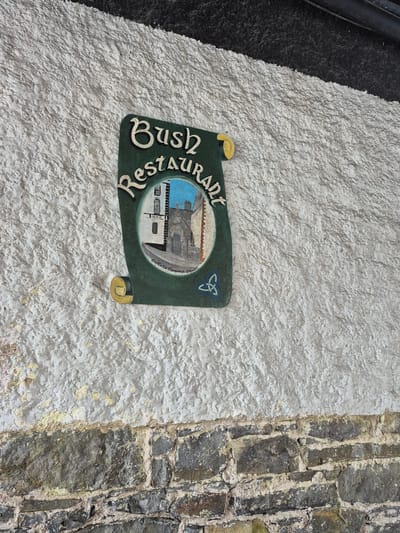 The image shows a sign for "Bush Restaurant" mounted on a textured white wall. The sign is green and scroll-shaped, with cream-colored lettering. Within the sign is a circular painting depicting a street scene with buildings and a sky. Below the painting, text reads "Custello Memorial Chapel". A Celtic knot symbol is also present on the sign. The wall is made of rough plaster, with some areas of peeling paint. Below the white wall is a section of dark stone masonry. The sign appears to be located outdoors, possibly on the side of a building. The city of Carrick-on-Shannon, Ireland, is mentioned in relation to the location.
