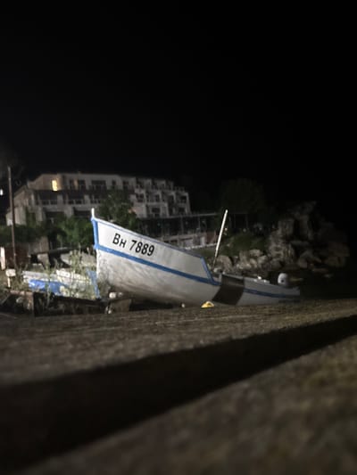 At night in Tyulenovo, Bulgaria, a white fishing boat with blue trim is beached on the shore. The boat has the registration number "BH 7889" painted on its side. Behind the boat, a multi-story building with illuminated windows is visible, suggesting it is a hotel or residential complex. The surrounding environment includes rocks, vegetation, and dark sky. The foreground is a blurred, dark surface. There are no people visible in the image.