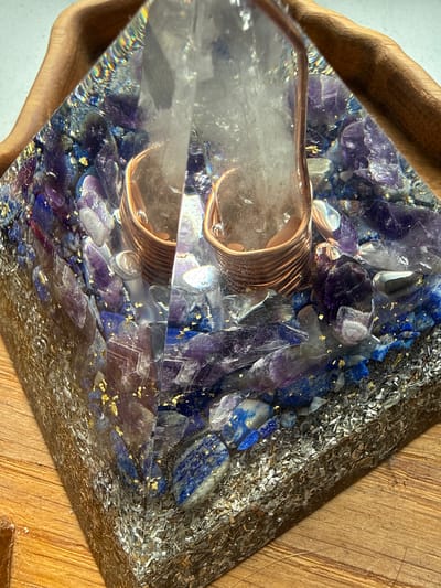An orgonite pyramid with amethyst and lapis lazuli crystals is displayed on a wooden surface. The pyramid is partially obscured by a curved piece of wood.