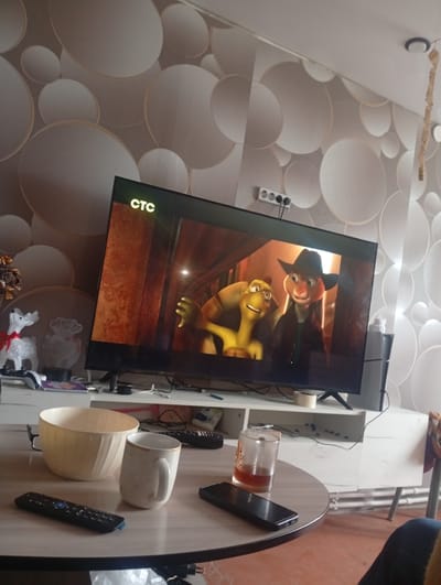 The image shows an indoor scene, likely a living room, with a large flat-screen television displaying a cartoon. The television is positioned on a white media console. The wall behind the television has a textured wallpaper with a pattern of overlapping circles. On the media console, to the left of the TV, are several decorative items, including a white, sparkly reindeer with a red ribbon and another white, silhouetted decoration. A game controller is also visible on the console.

In the foreground, a round table holds several objects. A large, light-colored bowl is prominent, accompanied by a white mug with a textured pattern and a golden rim. Two black remote controls are placed on the table, one near the bowl and mug, and another closer to the edge. A smartphone and a glass with a dark amber liquid are also on the table.

The overall impression is one of a cozy, perhaps slightly cluttered, domestic setting. The lighting suggests it is daytime, with artificial light from the TV and ambient room light. No individuals are clearly visible in the scene.

It is not possible to determine the specific location within Khomutovo, Russia, based on the visual information provided in the image.