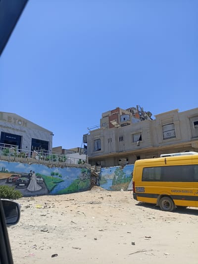 This media file captures a street scene under a clear blue sky, suggesting a bright, sunny day. The foreground is dominated by a sandy, debris-strewn lot, indicative of a somewhat undeveloped or possibly damaged area. A yellow bus with tinted windows is parked on the right side of the frame, displaying the text "KARDASH BUSES CO." along its side, along with two phone numbers: "0599 331382" and "0599 770252".
In the background, a mural adorns a wall, depicting a wedding scene with a bride and groom standing next to cars, a river, and a small house with trees. To the left of the mural is a modern building with large windows and the prominent text "LETON" above an entrance. Arabic text, "قاعة ليتون للمناسي" (Leton Hall for Occasions) and "بسم الله ماشاء الله" (In the name of Allah, what Allah has willed), is also visible on this building.
Further to the right, another building is under construction or appears to be damaged, with exposed concrete and some materials on its roof. The overall impression is a mixed environment with elements of commerce (the event hall and bus company) alongside signs of construction or disrepair, set against a backdrop of a vibrant mural. The presence of the wedding mural might suggest a place for celebrations, contrasting with the rougher elements of the surroundings.