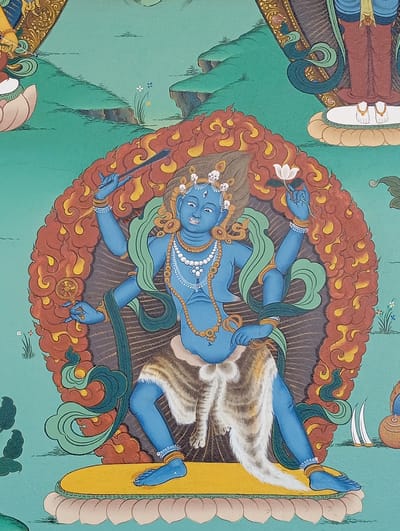 This is a detail of a painting depicting a blue-skinned, four-armed deity, possibly a Buddhist figure. The deity stands on a yellow lotus platform, positioned against a green backdrop with stylized landscape elements. A fiery, orange-red aureole surrounds the deity. The figure wears a skirt made of animal fur or skin and is adorned with jewelry like necklaces, armlets, and anklets. The deity holds various objects in each hand, including a staff-like object and a white lotus flower. The style is typical of Tibetan Buddhist art.
The image likely comes from Lumbini Sanskritik, Nepal, as it is a major Buddhist pilgrimage site.