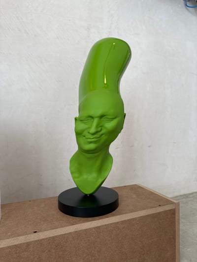 A bright lime green sculpture is positioned centrally on a light brown cuboid display stand, against a textured light gray wall, in San José del Cabo, Mexico. The sculpture consists of a human-like head with closed eyes and a subtle smile, rendered in a matte lime green finish. Extending vertically from the top of the head is an elongated, curved, tubular shape, finished in a glossy, reflective lime green. The sculpture rests on a circular, flat, matte black base, which sits atop the light brown stand. The stand appears to be constructed from particle board or MDF, with visible screw holes on its upper surface. A light gray floor is visible to the left foreground. Lighting creates reflections on the glossy upper portion of the sculpture and casts subtle shadows.