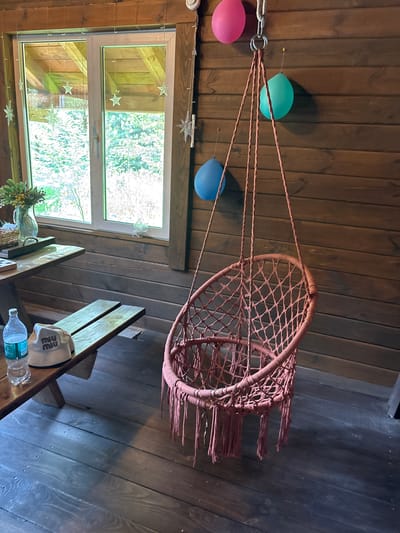 The image shows an indoor wooden room, possibly a porch or a cozy cabin, in Bol'shaya Rechka, Russia. The walls and floor are made of dark wooden planks. In the center of the image, a pink macrame hanging chair with fringe detailing is suspended from the ceiling by ropes. To the left, a wooden table and bench are set up. On the table, there is a glass vase with greenery, a woven basket, and what appears to be a chessboard. A plastic water bottle and a light-colored hat with a logo are placed on the bench. A large window on the left reveals a view of trees and greenery outside. Several colorful balloons—pink, turquoise, and blue—are attached to the wall and ceiling with pins, suggesting a festive occasion. The lighting appears to be natural, coming from the window and possibly an unseen light source.