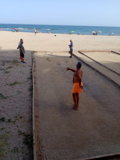 The image shows a beach scene in Águilas, Spain, with people playing boules on a sand court. In the foreground, a shirtless man wearing orange shorts and flip-flops is mid-throw, his arm extended and eyes focused on the boules court. Two other men are standing to the left, observing the game. The boules court is a rectangular area of compacted sand, bordered by wooden planks. Beyond the court, a sandy beach stretches towards the Mediterranean Sea, where several beach umbrellas and sunbathers are visible. The sky is clear and blue, and the sea is calm. The overall atmosphere suggests a relaxed, recreational activity on a sunny day.