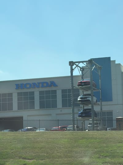 This is a wide, slightly blurred outdoor photograph of a Honda dealership. The building has a light-colored facade with a large blue panel on the right side. The word "HONDA" is displayed in large blue letters above a row of windows. To the right of the building, a car elevator holds three cars stacked vertically: a red car on top, a white car in the middle, and a dark blue car on the bottom. In front of the building, a black metal fence encloses a grassy area with several cars visible behind it. The sky is clear and blue. The location is a Honda dealership in Largo, United States of America.