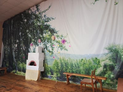 The image depicts a stage set with a backdrop of a natural landscape featuring trees and a sky. In the foreground, a white, multi-layered structure resembling a stove is positioned on the left side. To the right of the stove, a wooden bench is visible, with a wooden chair placed in front of it. The floor is made of wooden planks. The setting appears to be indoors, possibly a community center or a school, as indicated by the suspended ceiling tiles at the top of the image. There are no people or animals present in the scene. The backdrop is adorned with artificial flowers and star-shaped decorations. The overall scene is a static representation of a rustic outdoor environment. The location in the city of Gaynyamak, Russia, is a potential context for this stage set, suggesting a performance or an exhibition related to local culture or nature.