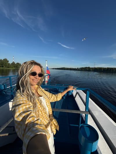A woman is depicted on the bow of a passenger boat, navigating a wide body of water, described as being in the city of Dumino, Russia, under a clear blue sky with sparse cirrus clouds. The woman, with long blonde and white dreadlocks, is wearing dark oval sunglasses, a yellow and white checkered long-sleeve shirt over a white top, and is smiling towards the camera. Her right hand rests on the boat's blue metal railing. Behind her, a Russian flag is visible on a pole, with partially obscured Cyrillic text below it. The boat features blue railings, deck elements, and a blue cylindrical container on its white deck structure. The water is calm, exhibiting dark blue hues and surface ripples. On the distant banks, dense green treelines are visible. A prominent red and white striped lattice tower stands on the left bank, and another less distinct tower is visible on the far right bank. Several seagulls are observed flying low over the water and resting on its surface. The scene is illuminated by direct sunlight, casting shadows from the boat's structure.