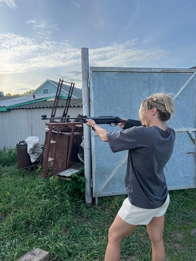 A person with blonde hair in a bun, wearing a gray t-shirt and white shorts, is holding a black shotgun in their hands. The person is standing outdoors in a grassy area with a metal structure and a building in the background. The sky is visible with clouds. The location is Слобода, Russia.