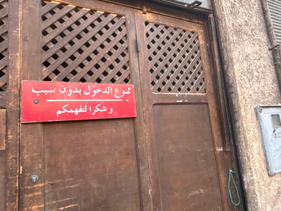 The image shows a wooden double door with a red sign affixed to the left door panel. The sign has white Arabic text, which translates to "No entry without invitation and thank you for your understanding." The doors have a diagonal slatted design. To the right of the doors is a textured concrete wall with what appears to be an electrical meter box attached. A green string is tied to the door and hangs loosely. The scene is likely an entrance to a private property or establishment in Victoria, Morocco.